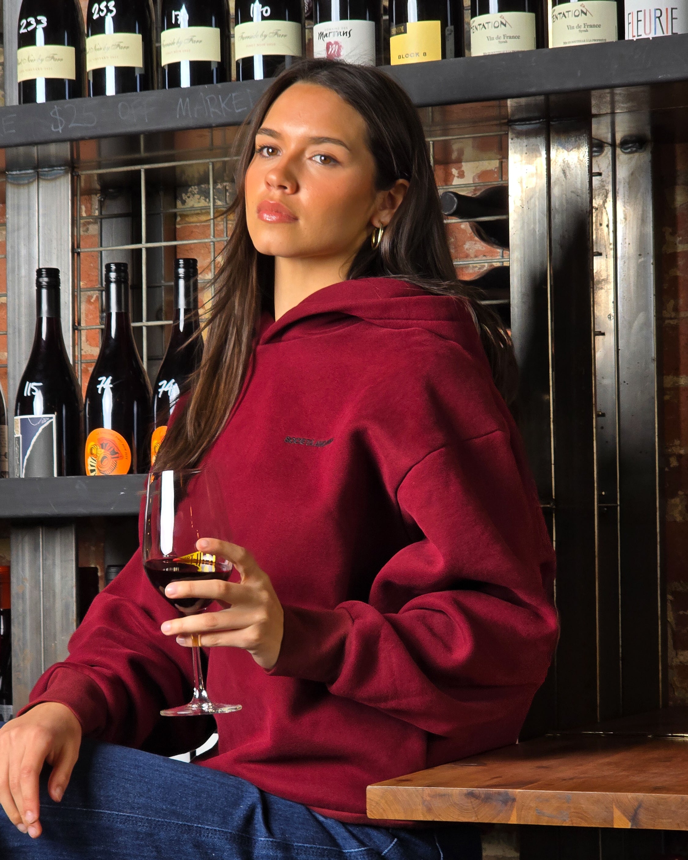 Woman in a red hoodie holding a glass of wine in front of wine bottles.