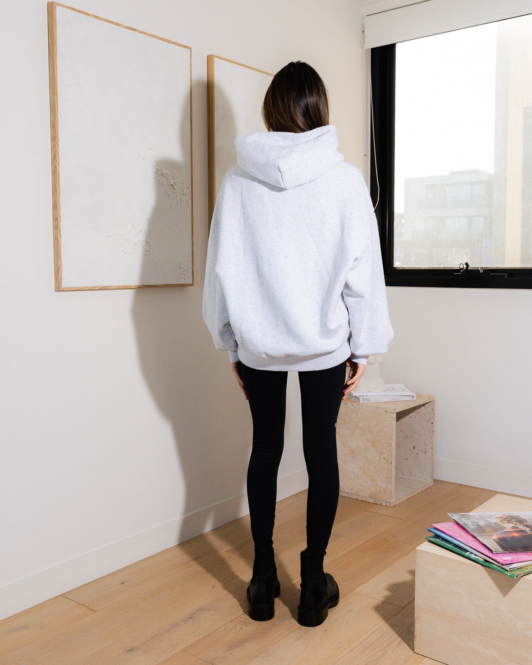 Woman wearing a heather grey hoodie with ANONIMA branding in a room with light-colored walls and wooden floor.