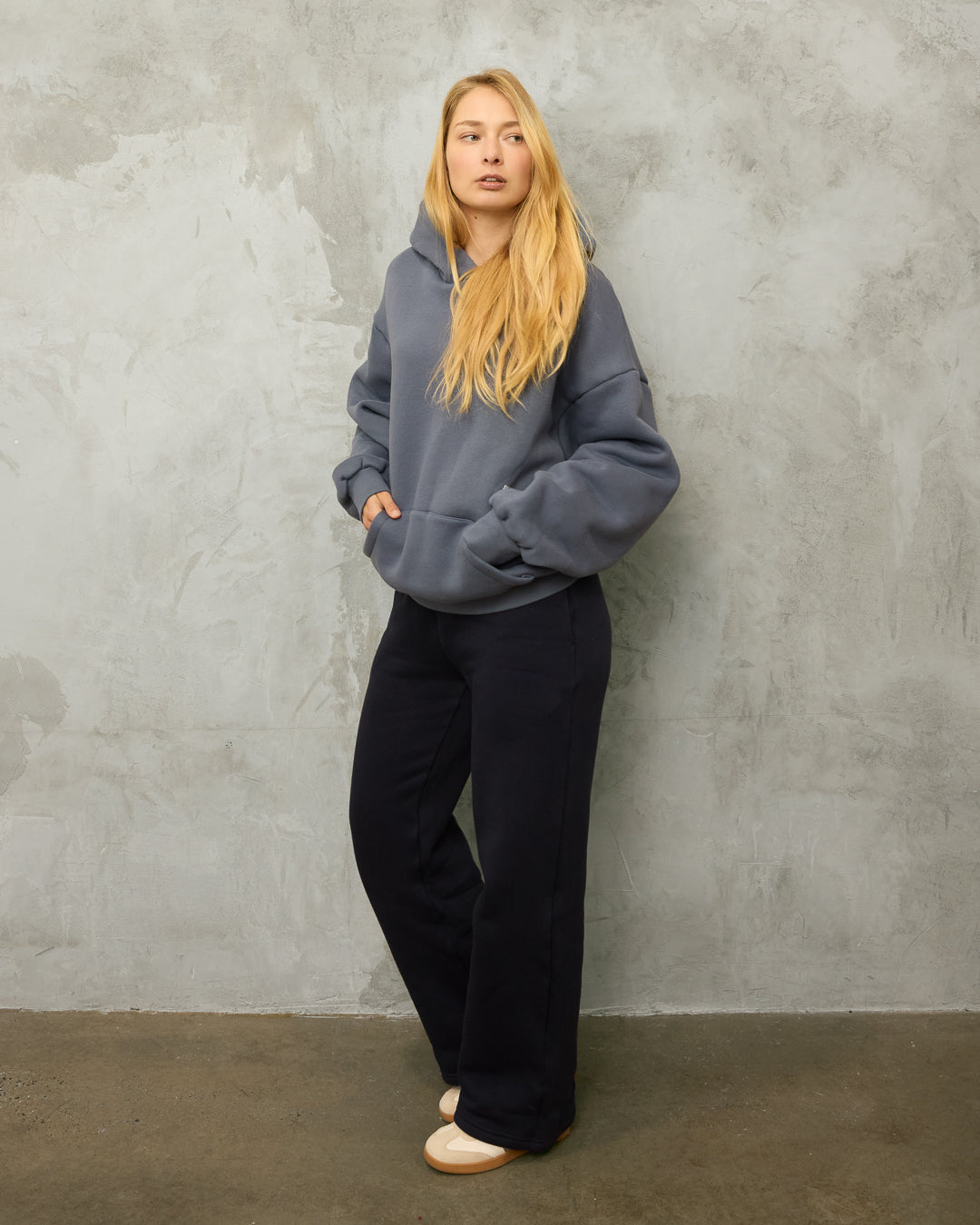 Person wearing a gray hoodie and midnight blue pants against a textured gray wall.