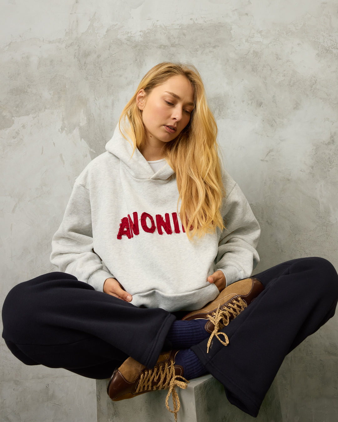 Person wearing a hoodie with 'ANONIMA' text, sitting on a concrete surface with midnight blue track pants.