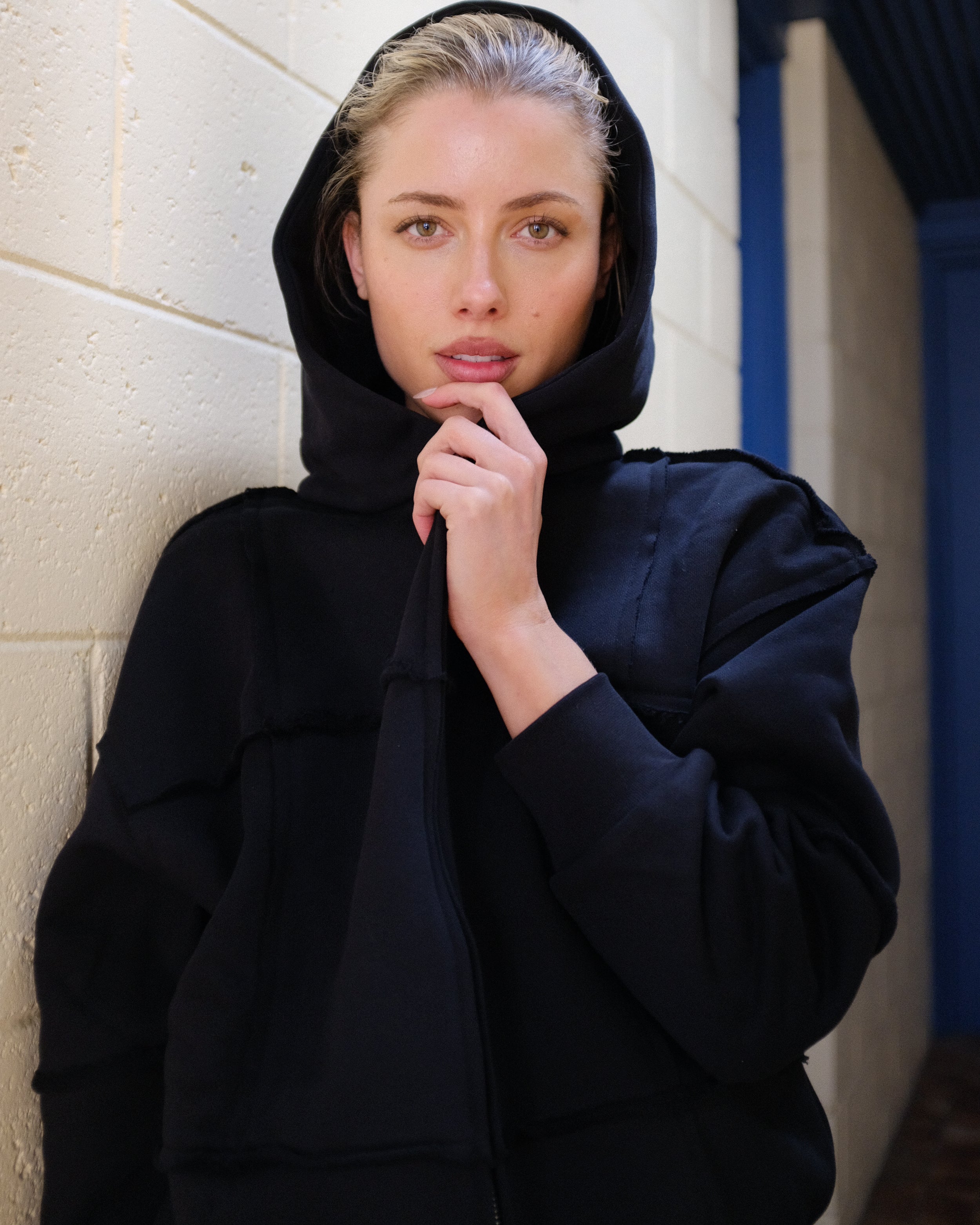 Woman wearing a black hooded outfit against a light-colored wall.