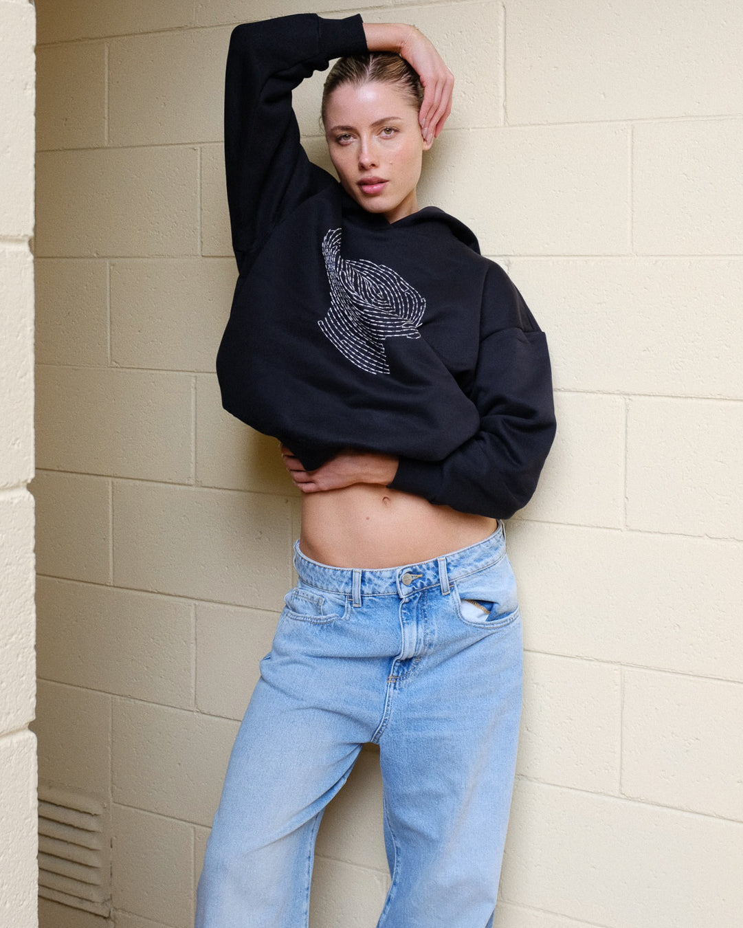 Person wearing a black hoodie with a graphic design and light blue jeans against a beige wall.