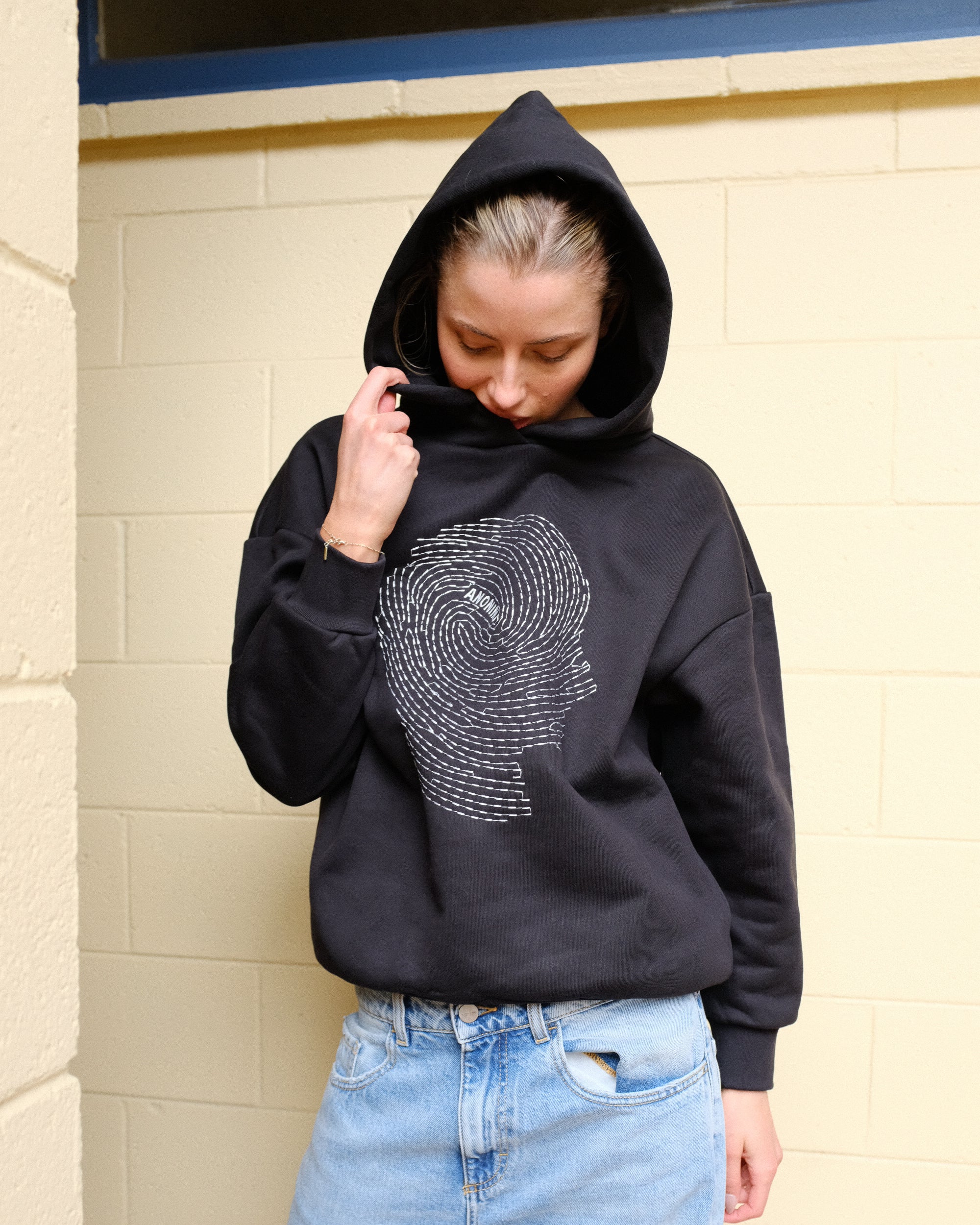 Person wearing a black hoodie with a white graphic design, standing against a beige wall.