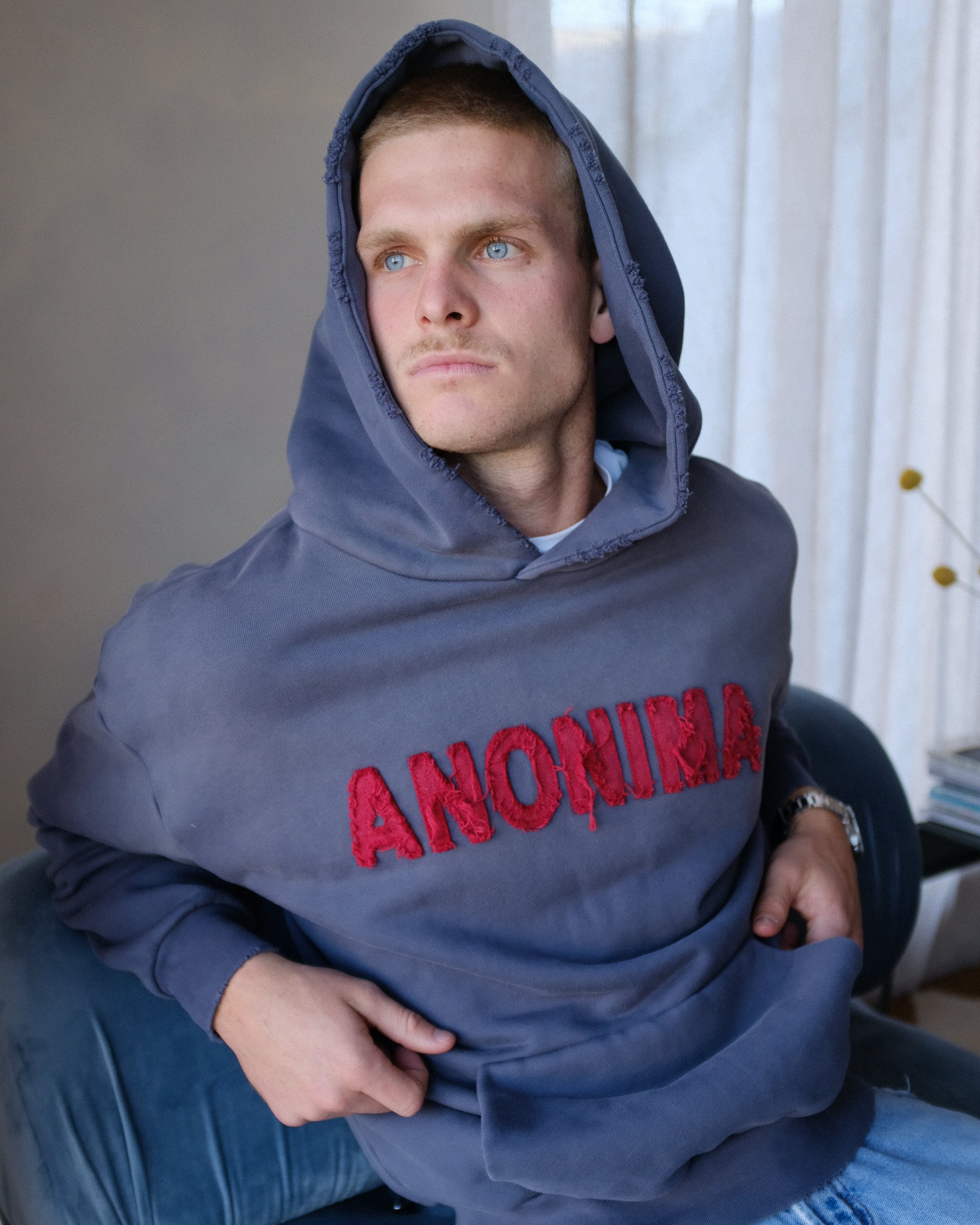 Man wearing a blue hoodie with 'ANONIMA' in red text, sitting indoors.