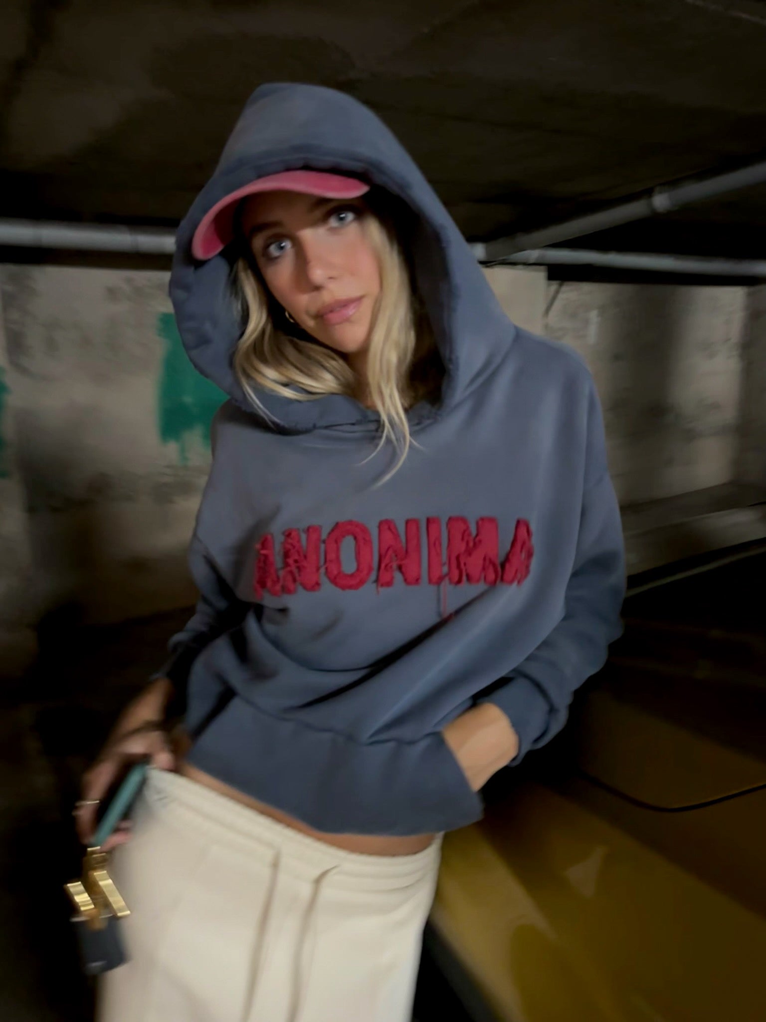 Person wearing a blue hoodie with 'ANONIMA' text, standing indoors.