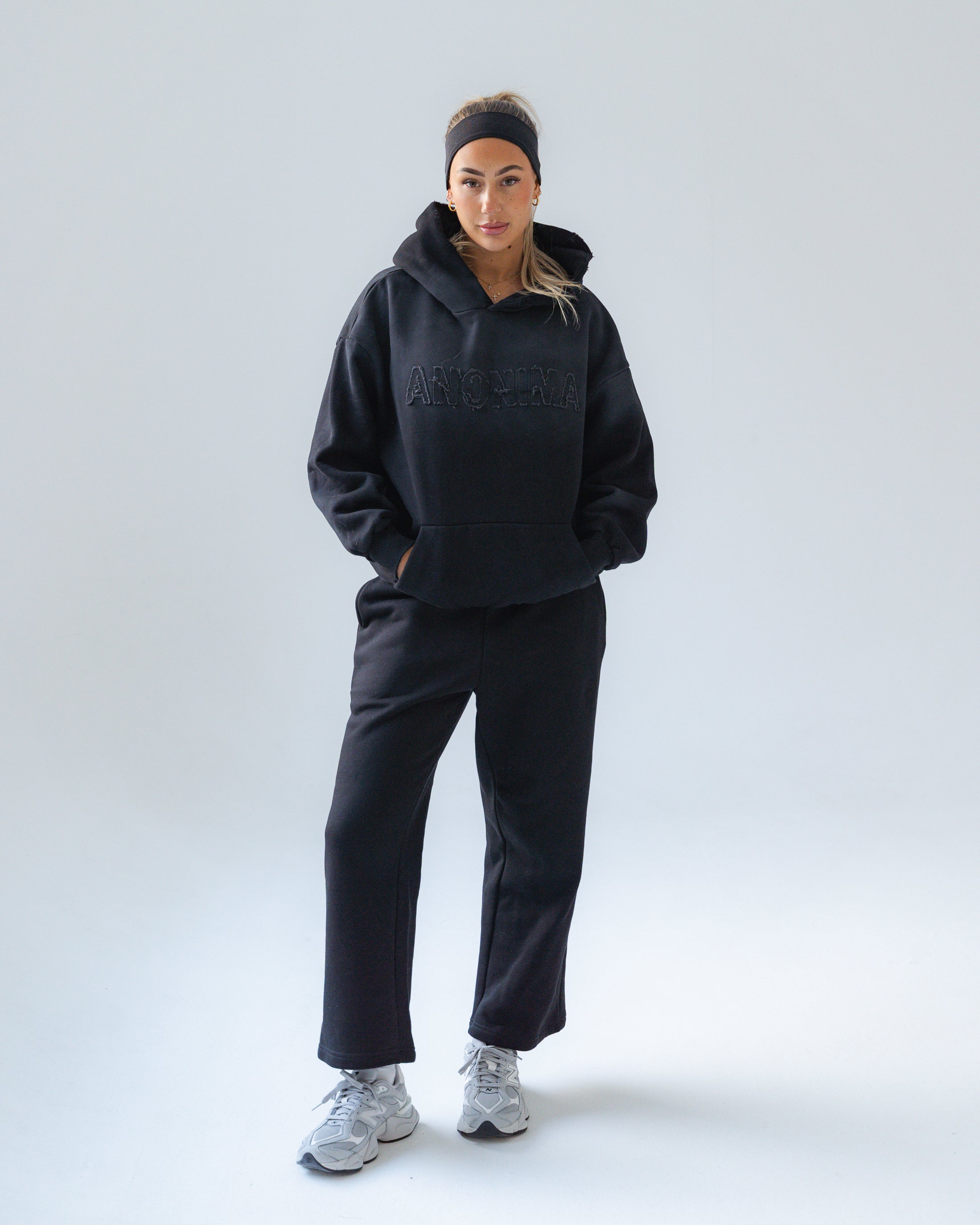 Person wearing a black hoodie and pants with 'ANONIMA' text on a white background