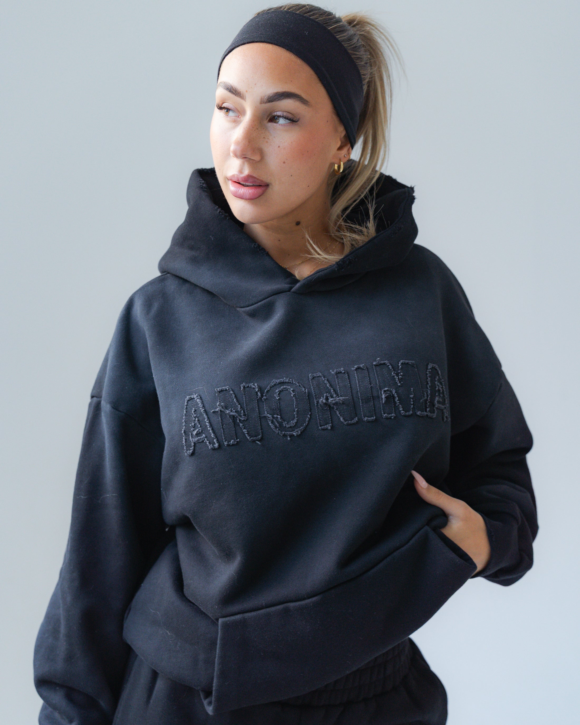 Person wearing a black hoodie with 'ANONIMA' text on a plain background
