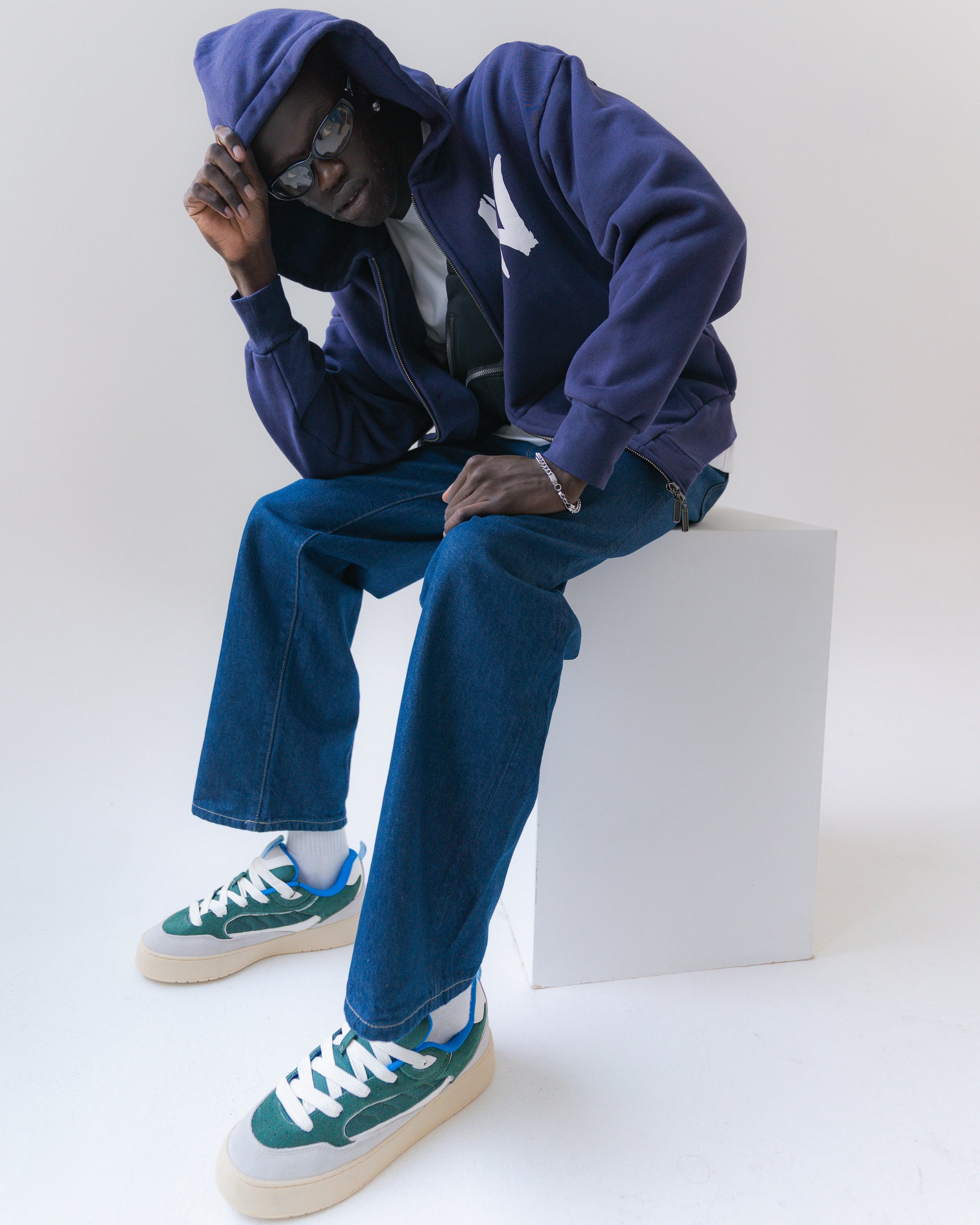 Person wearing a blue hoodie, jeans, and green sneakers sitting on a white block.