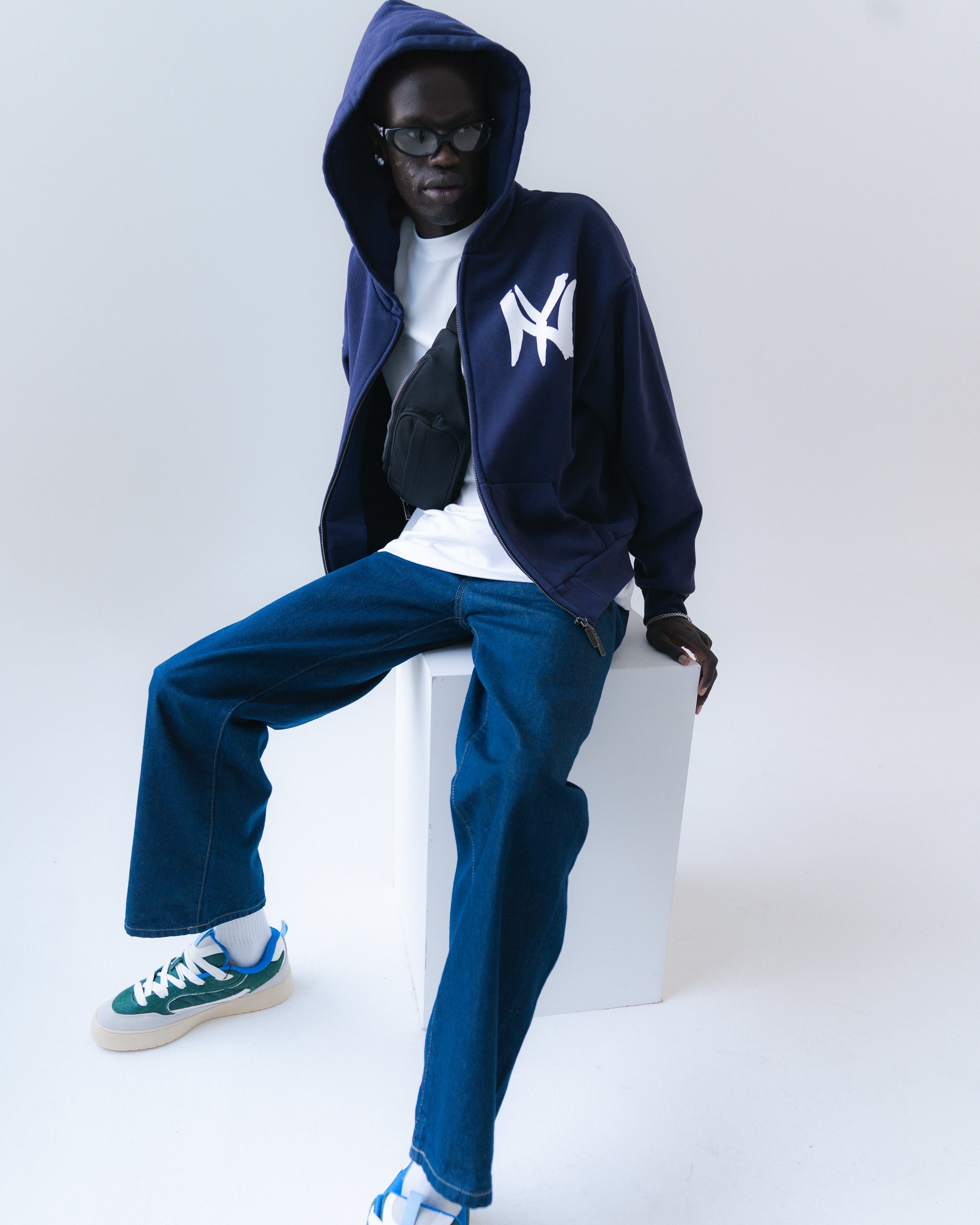 Person wearing a navy hoodie with a logo, white shirt, blue jeans, and sneakers on a white background
