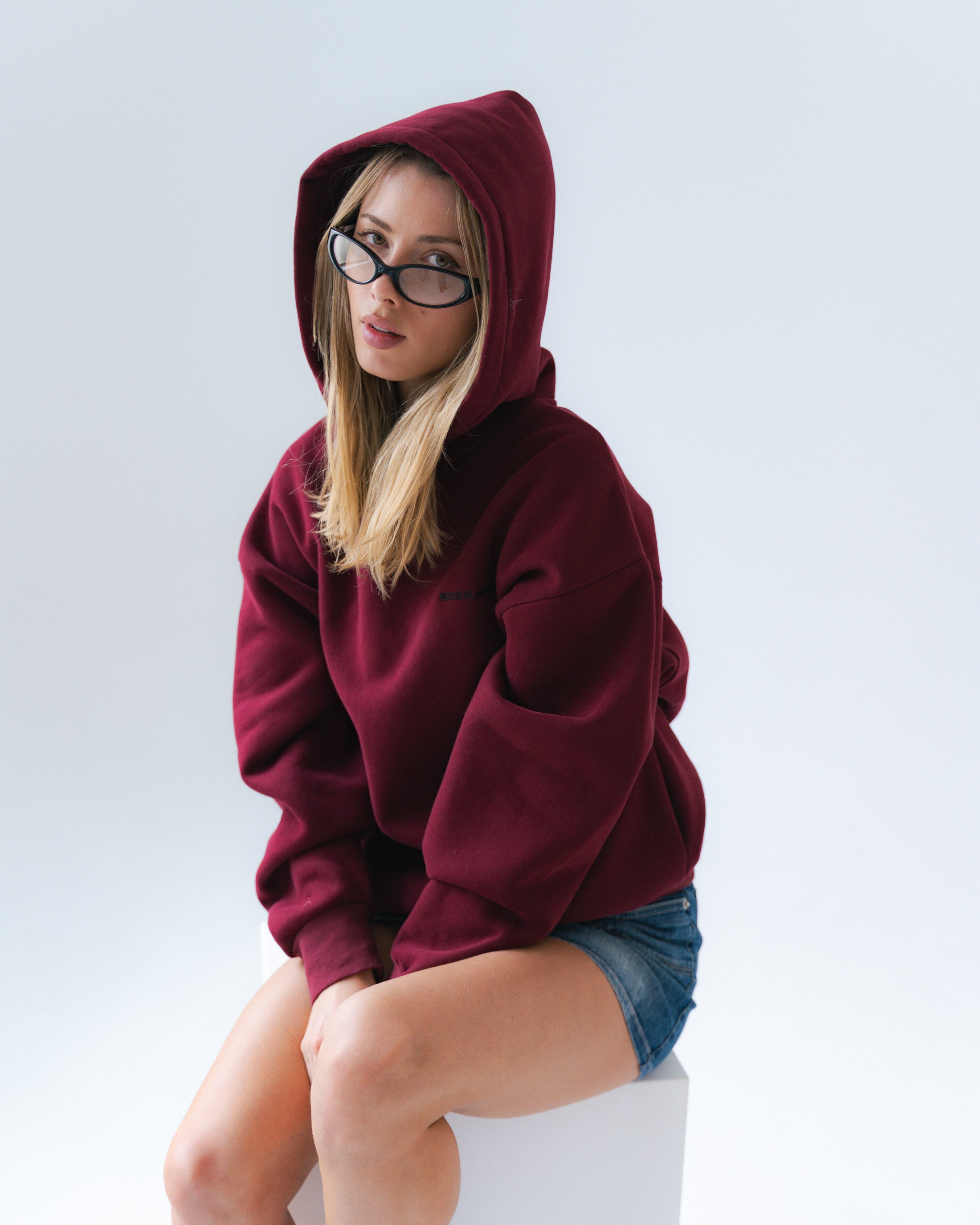 Woman wearing a maroon hoodie and denim shorts sitting on a white box against a light gray background