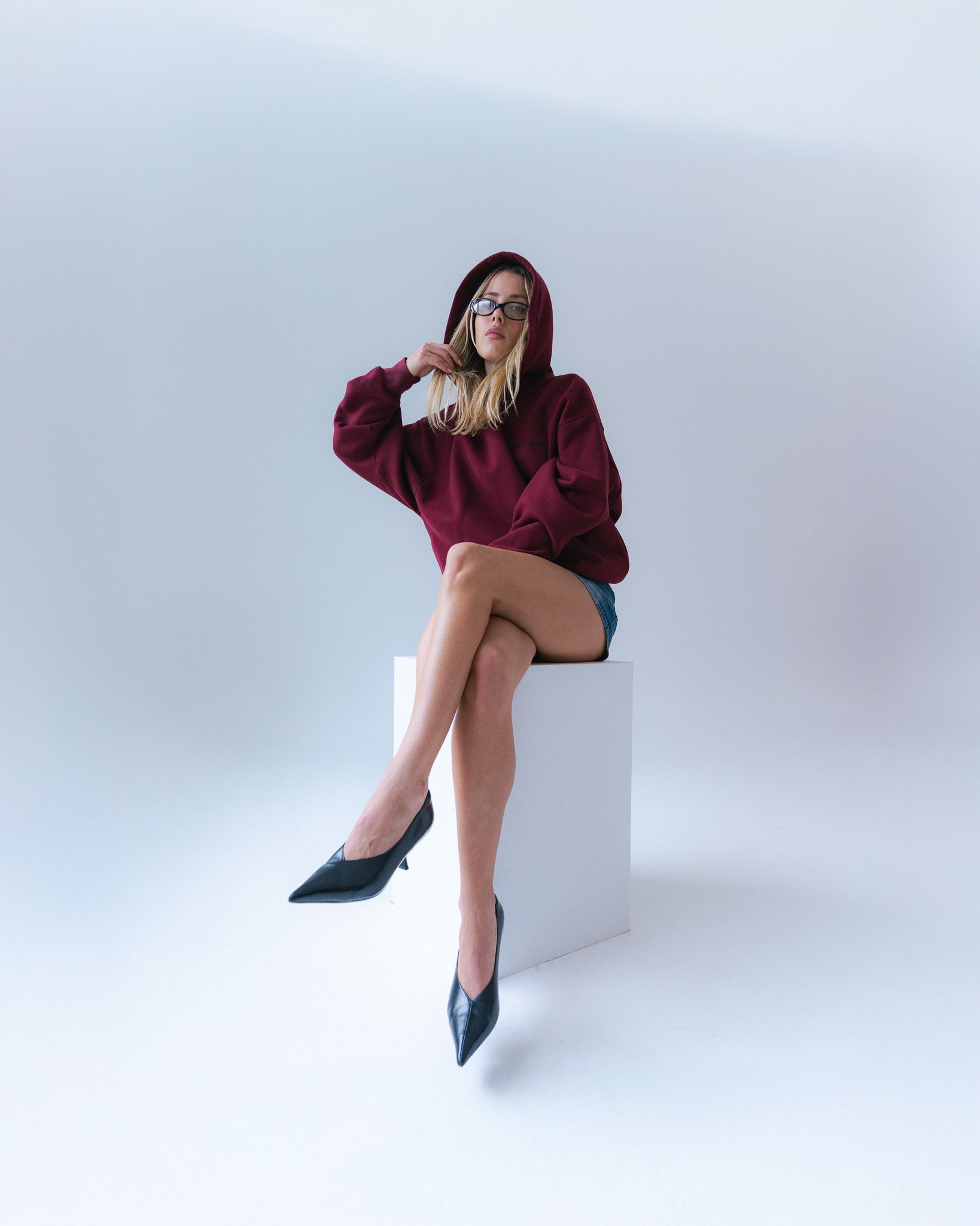 Woman wearing a burgundy hoodie and denim shorts sitting on a white cube against a light gray background